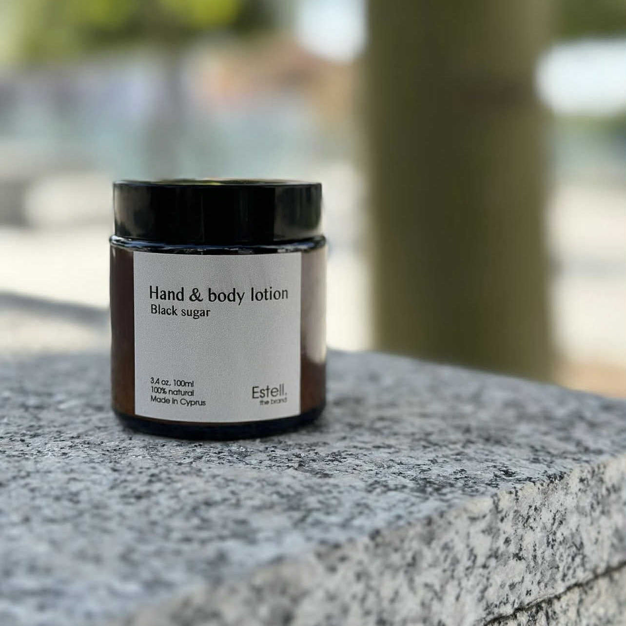 Hand and body lotion jar labeled 'Black sugar' by Estelle on a stone surface with a blurred natural background.