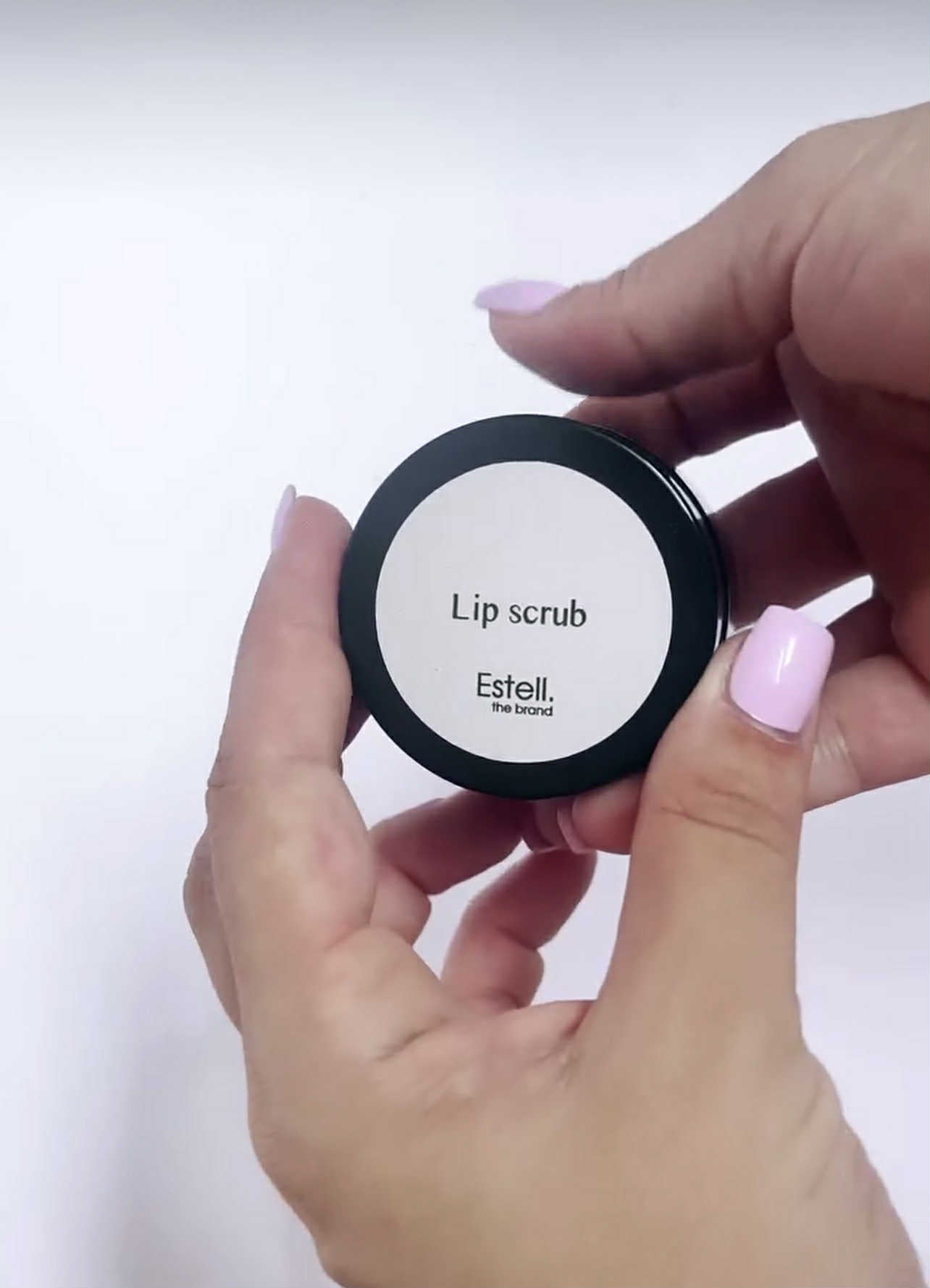 Lip scrub container labeled 'Estell' held by a hand with pink nail polish on a white background